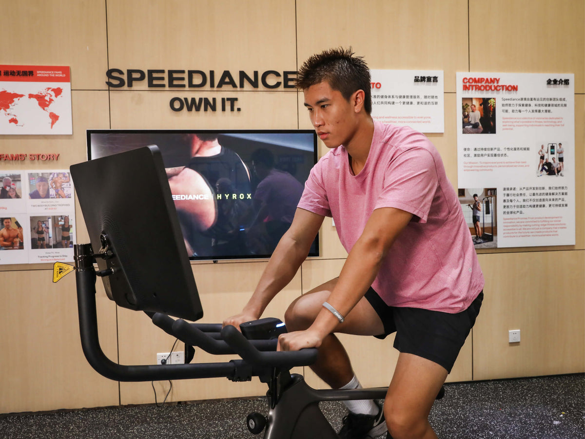 Eine Person, die in einem Raum auf einem Heimtrainer Fahrrad fährt. An der Wand hängt ein Schild mit der Aufschrift "Speediance".